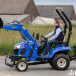 Iseki TXGS 24 met voorlader en gewichtsbak achterop, bestuurder op de tractor langs een weg