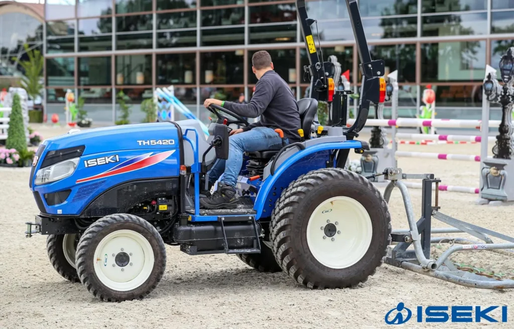 Iseki TH 5420 met veiligheidsbeugel en een zandhark achterop, met bestuurder