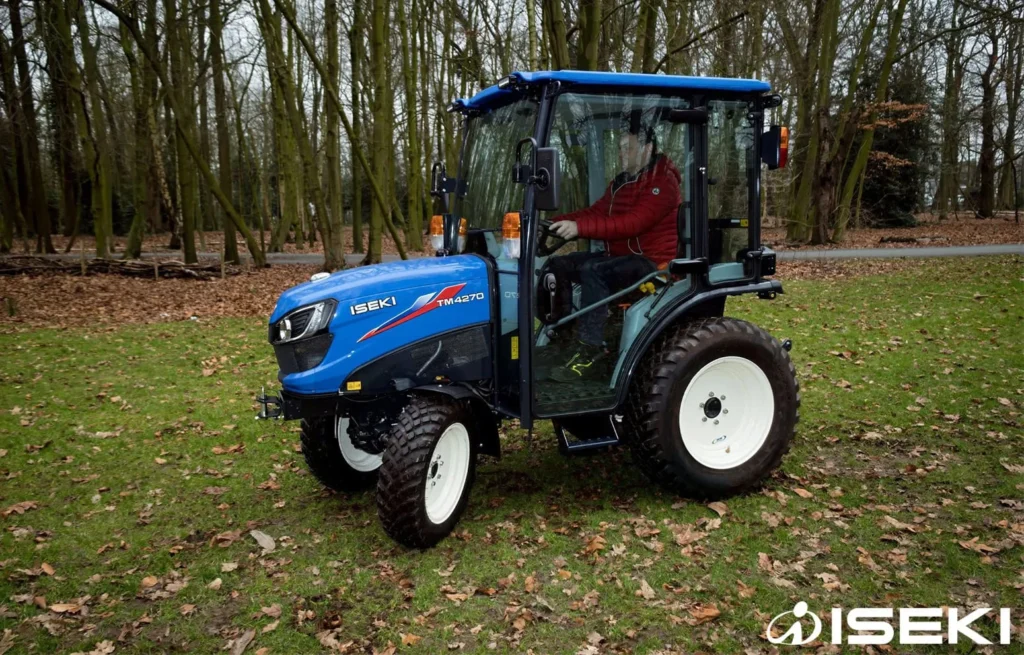 Bestuurder in rode jas in de cabine van de Iseki TM 4270 op een grasveld met bladeren