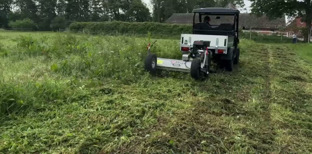 Maaier met eigen motor achter een terreinvoertuig in een weiland, met een gemaaide baan in het hoge gras