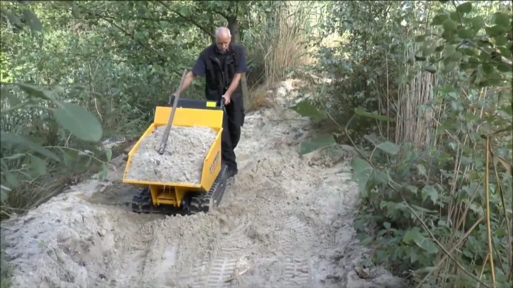 Rupsdumper met een bak zand op een smal pad van los zand tussen struiken